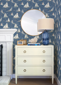 A white dresser with three drawers sits against Chesapeake Leeward Sailboat Wallpaper by York Wallcoverings. A round mirror, a blue glass lamp, coral decor, and framed photos rest on top. Part of a white fireplace is visible on the left.
