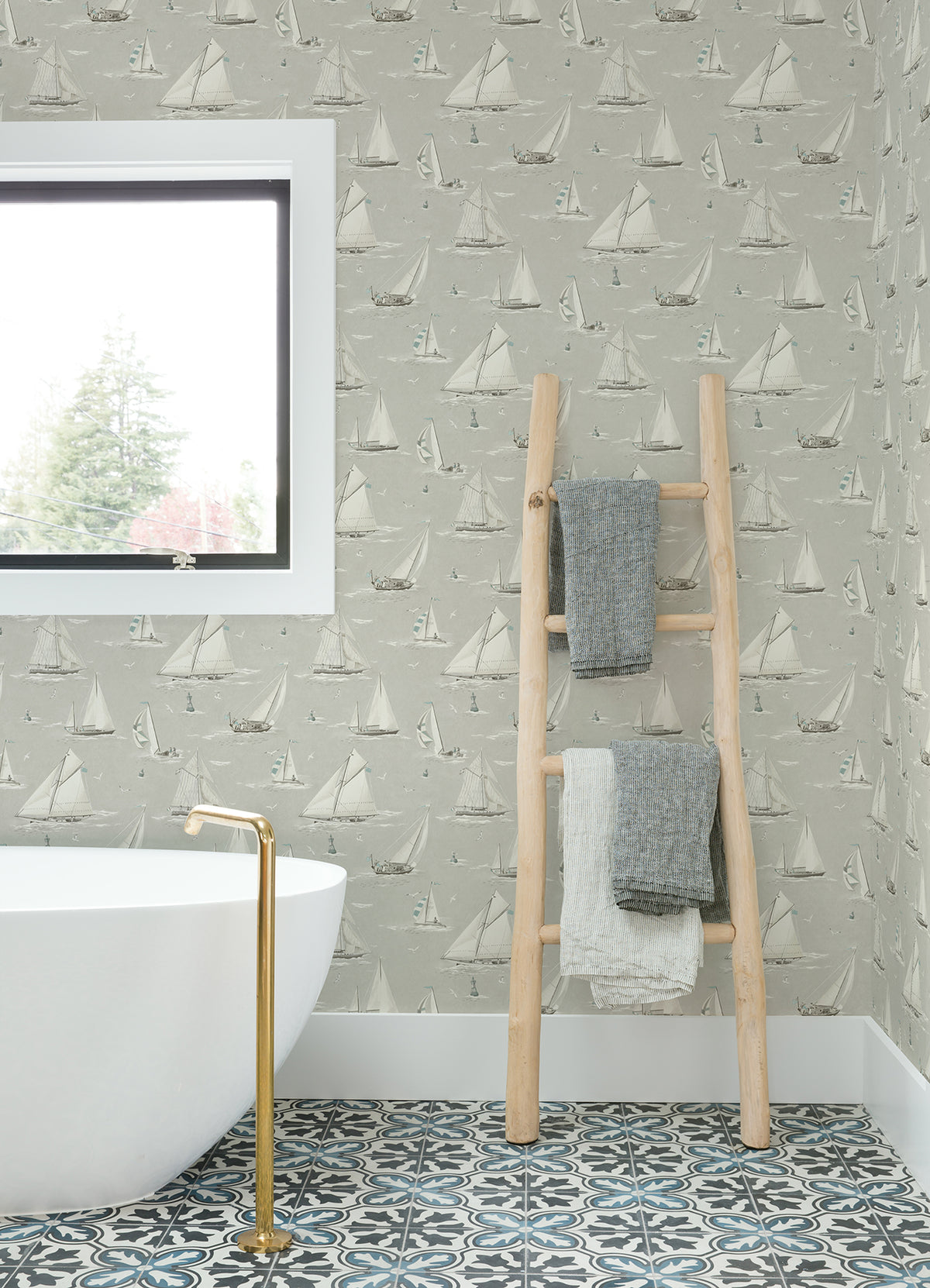 A bathroom featuring a white tub, gold faucet, patterned tile floor, window, and a wooden ladder holding three folded towels is adorned with York Wallcoverings’ Chesapeake Leeward Sailboat Wallpaper.