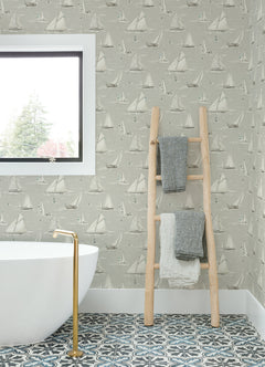 A bathroom featuring a white tub, gold faucet, patterned tile floor, window, and a wooden ladder holding three folded towels is adorned with York Wallcoverings’ Chesapeake Leeward Sailboat Wallpaper.