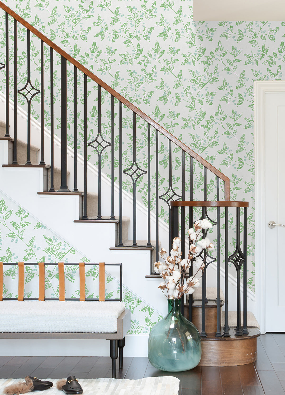 A stairway with black railings and wood handrails is next to a wall covered in Ardell Botanical Wallpaper by York Wallcoverings. Beside the stairs sits a bench with tan straps and a glass vase filled with cotton stems.