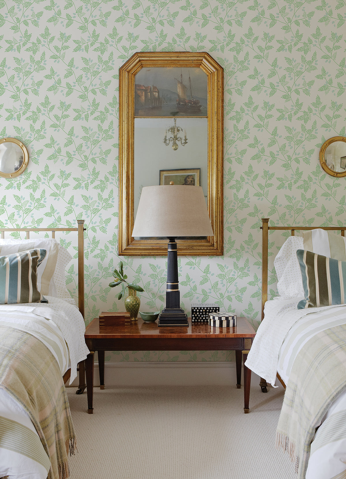 Two twin beds with striped pillows and plaid throws are separated by a wooden table topped with a lamp, small decor pieces, and a leafy plant. A large mirror with a gold frame hangs above the table on walls adorned with Ardell Botanical Wallpaper by York Wallcoverings.