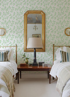 Two twin beds with striped pillows and plaid throws are separated by a wooden table topped with a lamp, small decor pieces, and a leafy plant. A large mirror with a gold frame hangs above the table on walls adorned with Ardell Botanical Wallpaper by York Wallcoverings.