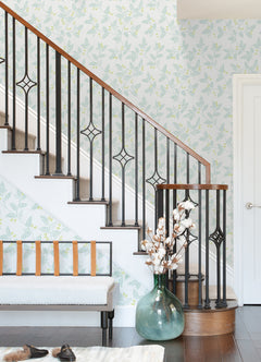 A staircase with black metal railings, Ardell Botanical Wallpaper by York Wallcoverings in a light floral pattern, a bench with leather straps, and a large green vase holding cotton stems are featured in a bright, modern entryway.