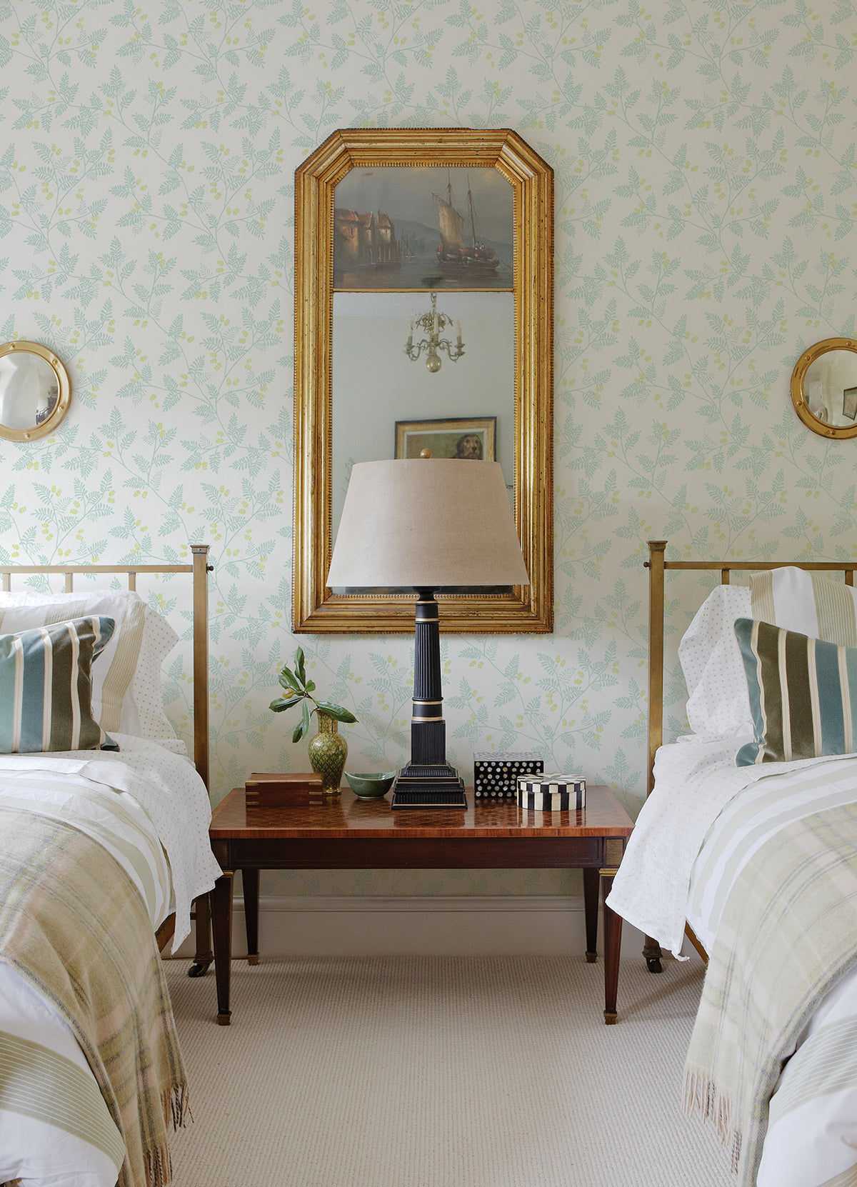 Two twin beds with striped pillows flank a wooden nightstand topped with a table lamp, small plant, and decor. Behind the beds, a large gold-framed mirror and round mirrors hang on York Wallcoverings’ Ardell Botanical Wallpaper.