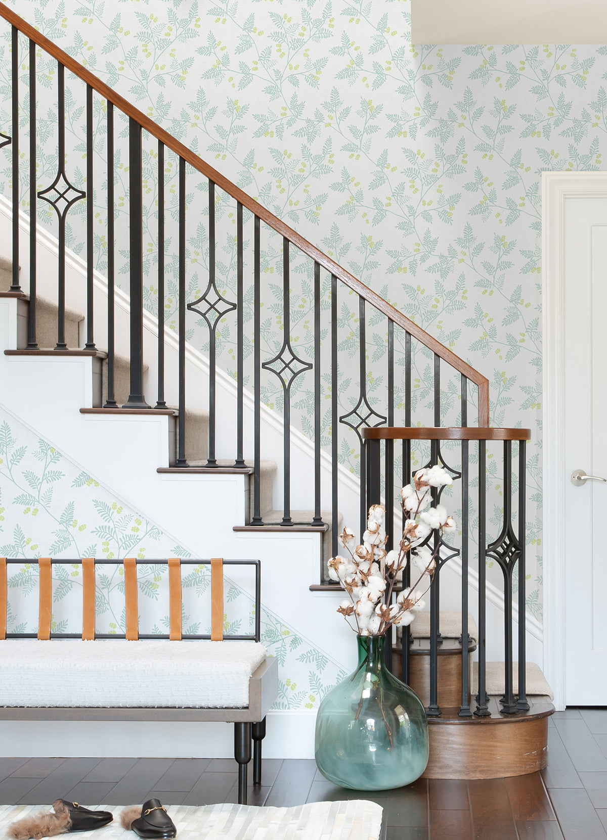 A staircase with black iron rails and a wooden handrail stands next to a bench with leather straps. Against the wall, covered in Ardell Botanical Wallpaper by York Wallcoverings, a large green vase filled with cotton stems sits on dark wood flooring.