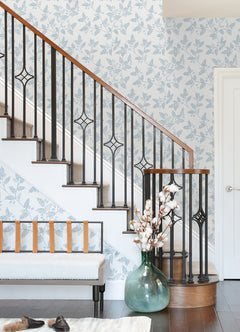 A curved staircase with black metal railings, Ardell Botanical Wallpaper by York Wallcoverings, a glass vase with cotton stems, a bench with tan straps, shoes on the floor, and a white door in the background.