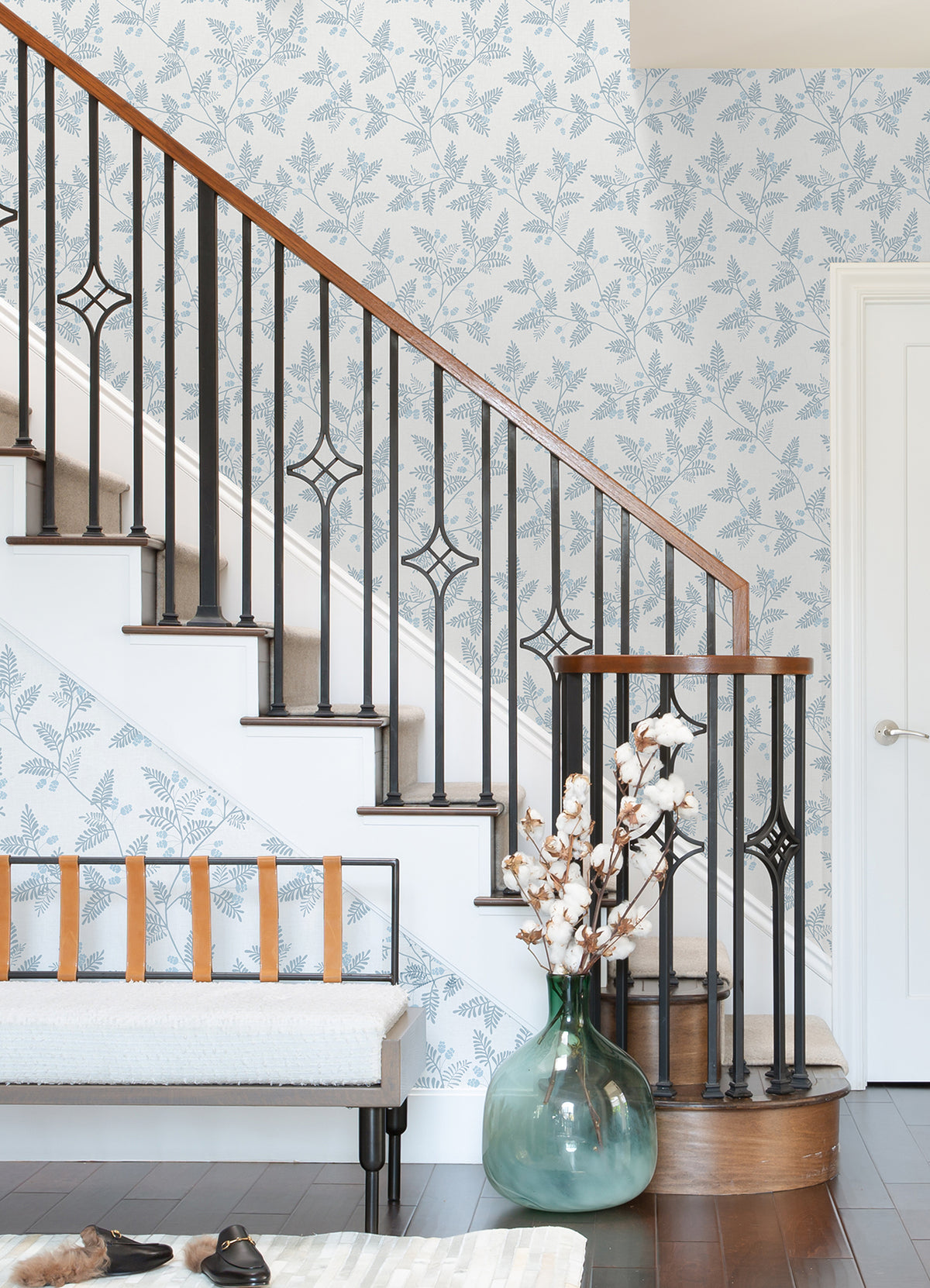A staircase with a black metal railing and wood handrail, Ardell Botanical Wallpaper by York Wallcoverings, a bench with leather straps, a large green vase with cotton stems, and brown shoes on a dark wood floor.