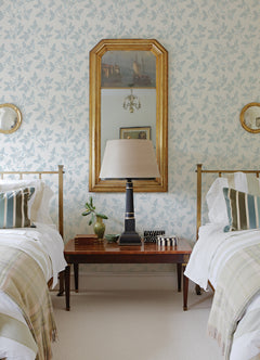 Two twin beds with white bedding flank a wooden nightstand topped by a lamp, plant, and books. Above the nightstand, a large gold-framed mirror and two round mirrors hang on York Wallcoverings’ Ardell Botanical Wallpaper.
