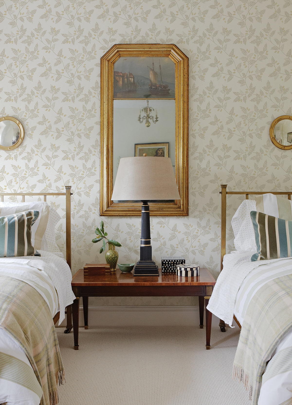 Two twin beds with striped pillows flank a wooden table holding a lamp, a plant, and decor; a gold-framed mirror and art hang above, reflecting a chandelier. The walls are adorned with Ardell Botanical Wallpaper by York Wallcoverings, featuring a subtle floral pattern.