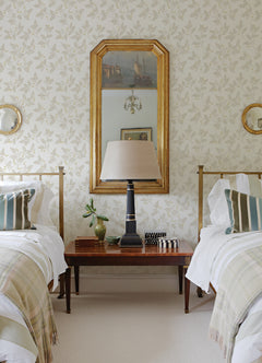 Two twin beds with striped pillows flank a wooden table holding a lamp, a plant, and decor; a gold-framed mirror and art hang above, reflecting a chandelier. The walls are adorned with Ardell Botanical Wallpaper by York Wallcoverings, featuring a subtle floral pattern.