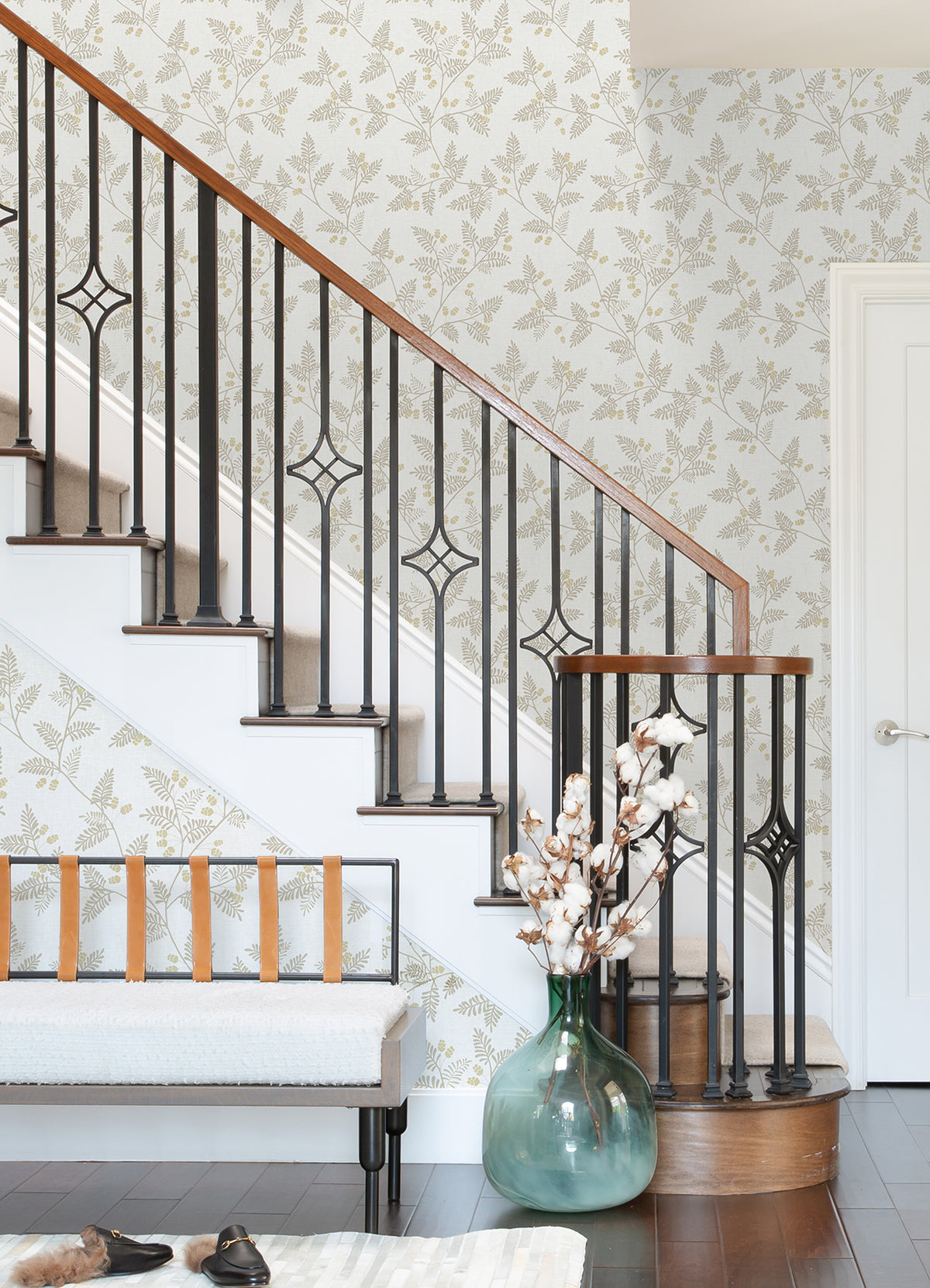 A staircase with a black metal railing and wooden handrail stands beside a white bench with leather straps. On the dark wood floor rests a large green vase filled with cotton stems. The wall behind is adorned with Ardell Botanical Wallpaper by York Wallcoverings, featuring a beautiful floral design.