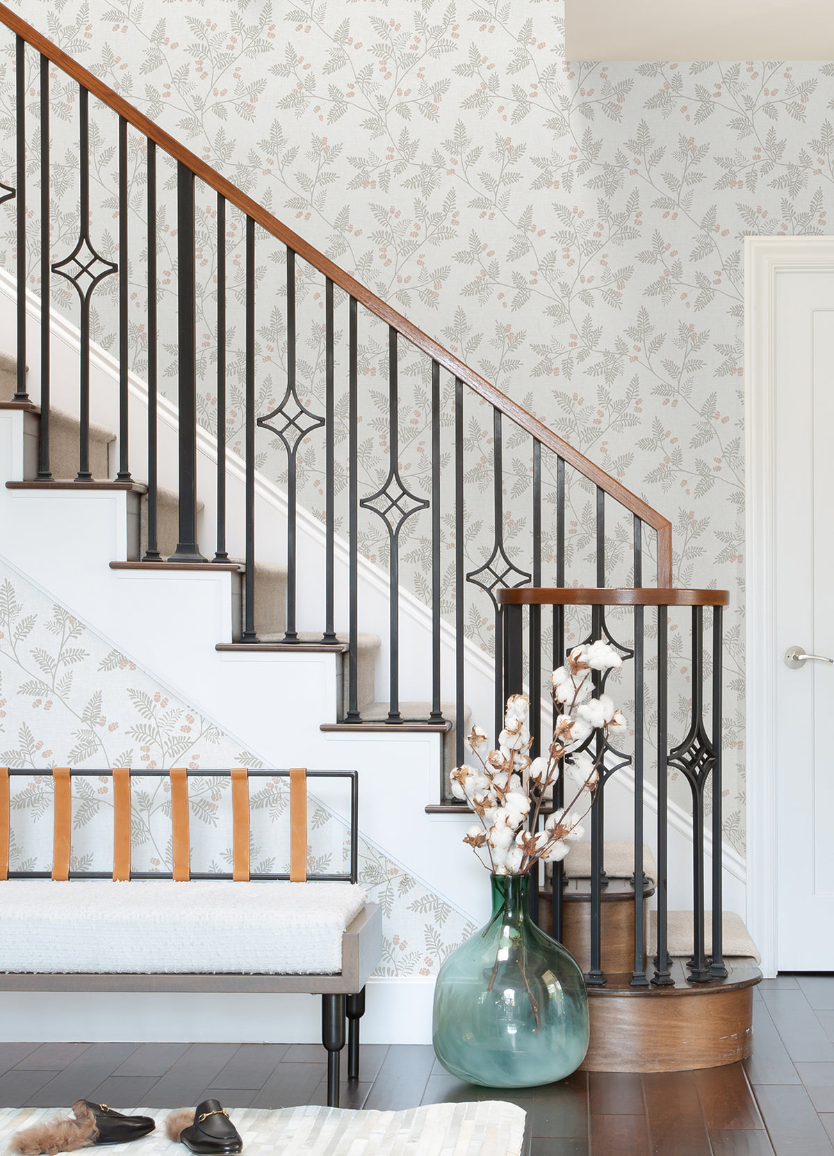 A staircase with black iron railings and a wooden handrail stands beside a bench with tan straps. A green vase filled with cotton stems rests on the dark wood floor, while the wall is adorned with Ardell Botanical Wallpaper by York Wallcoverings.