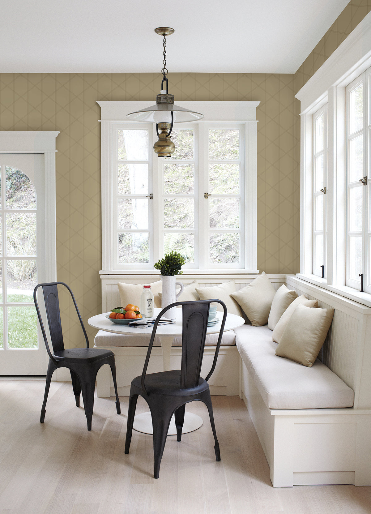 Bright breakfast nook featuring Walcott Stitched Trellis Wallpaper by York Wallcoverings, a round white table, black chairs, and a built-in white bench with beige pillows. Large windows fill the space with natural light, while fruit and coffee sit atop the table.