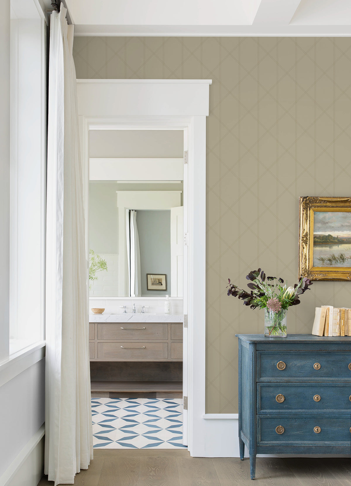A bright room featuring York Wallcoverings’ Walcott Stitched Trellis Wallpaper, a blue dresser, gold-framed painting, vase of flowers, white trim, and a view into a bathroom with a patterned tile floor and wooden vanity.