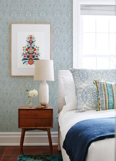 A mid-century nightstand with a lamp, a small vase of flowers, and framed art above sits next to a bed with blue and white pillows and a dark blue throw in a bright bedroom featuring Fernback Ornate Botanical Wallpaper by York Wallcoverings.