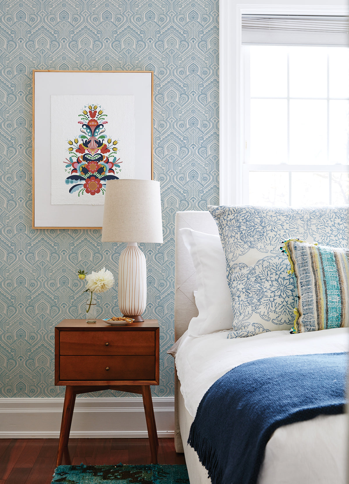 A cozy bedroom featuring a mid-century nightstand, a ceramic lamp, flowers, and a bed adorned with blue and white pillows and blanket. Fernback Ornate Botanical Wallpaper by York Wallcoverings adds pattern behind a framed artwork beside a large window.