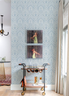 A bar cart with glasses and bottles stands against a wall adorned with York Wallcoverings’ Fernback Ornate Botanical Wallpaper. Two blurry portraits of a woman in a green dress hang above the cart, while sunlight streams in from a window with beige curtains on the right.
