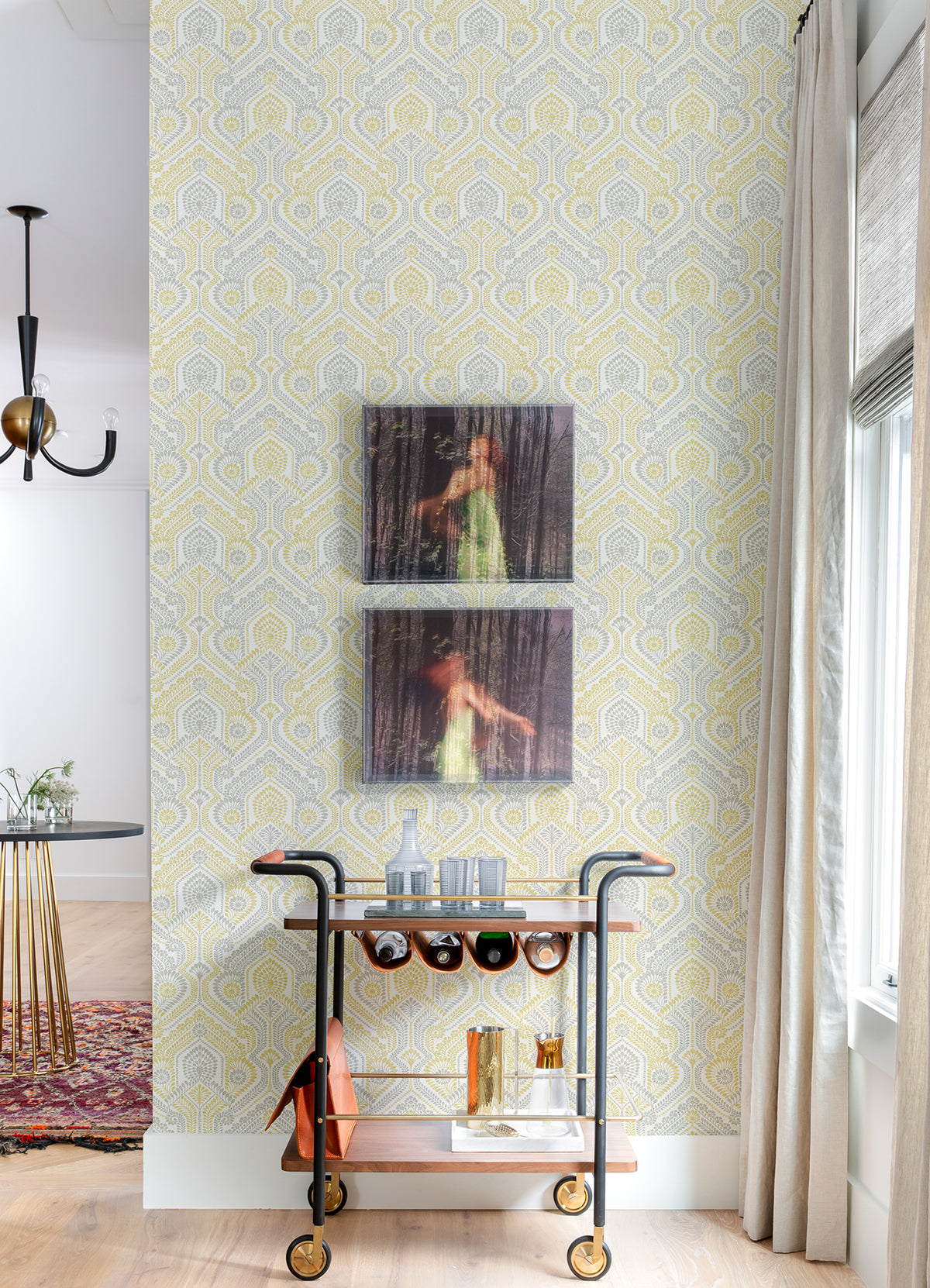 A bar cart with bottles and glasses stands against a wall adorned with York Wallcoverings’ Fernback Ornate Botanical Wallpaper, with two blurred photos of a woman in a green dress hanging above. A window with light curtains is to the right, and a dining area is visible to the left.