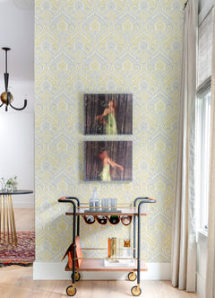 A bar cart with bottles and glasses stands against a wall adorned with York Wallcoverings’ Fernback Ornate Botanical Wallpaper, with two blurred photos of a woman in a green dress hanging above. A window with light curtains is to the right, and a dining area is visible to the left.