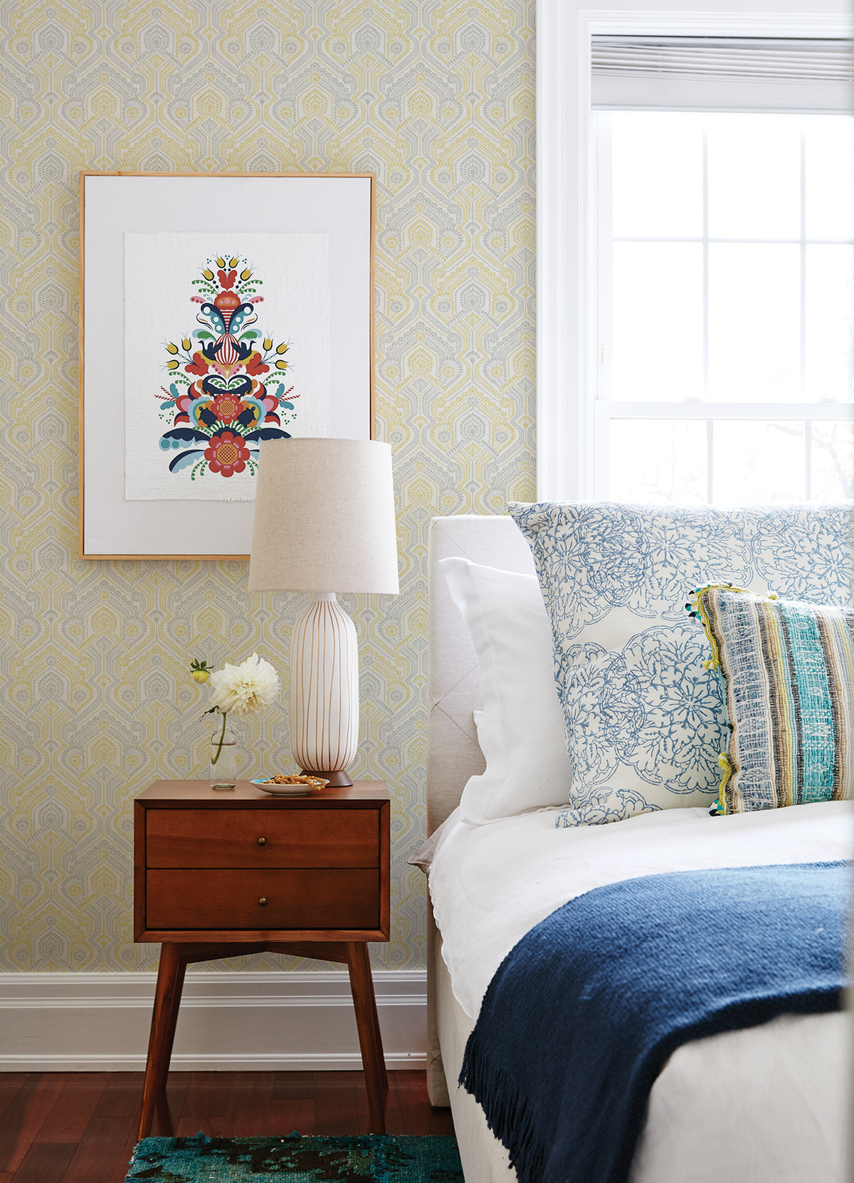 A cozy bedroom featuring a white bed with blue and patterned pillows, a blue blanket, a wooden nightstand topped with a lamp and flower vase, and framed floral artwork on York Wallcoverings’ Fernback Ornate Botanical Wallpaper beside a bright window.
