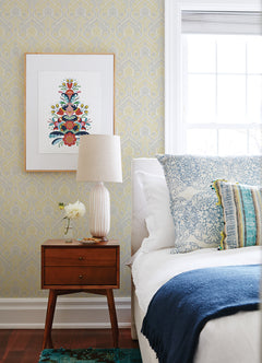 A cozy bedroom featuring a white bed with blue and patterned pillows, a blue blanket, a wooden nightstand topped with a lamp and flower vase, and framed floral artwork on York Wallcoverings’ Fernback Ornate Botanical Wallpaper beside a bright window.