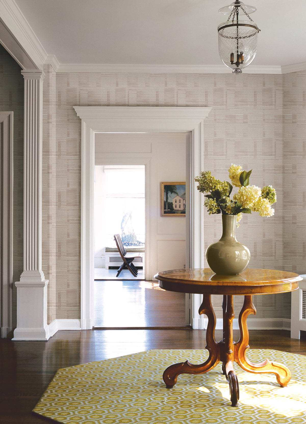 A bright hallway with columns, wooden floors, and a hanging light fixture features Callaway Woven Stripes Wallpaper by York Wallcoverings. A wooden table with a green vase of flowers sits on a yellow patterned rug, while through the doorway, a sunlit room with a chair is visible.