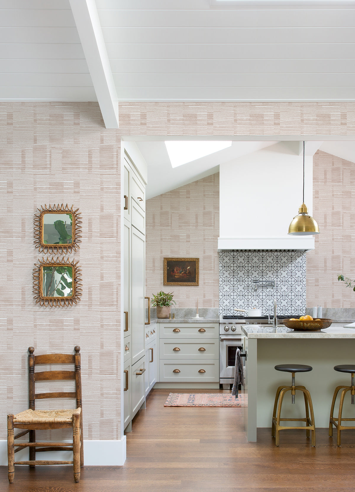 A bright kitchen features Callaway Woven Stripes Wallpaper by York Wallcoverings, a gold pendant light, a green island with stools, and a patterned backsplash. A wooden chair and framed art are displayed on the wall, while wood flooring adds warmth to the space.
