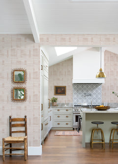 A bright kitchen features Callaway Woven Stripes Wallpaper by York Wallcoverings, a gold pendant light, a green island with stools, and a patterned backsplash. A wooden chair and framed art are displayed on the wall, while wood flooring adds warmth to the space.