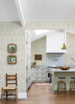 Bright kitchen featuring a patterned backsplash, white cabinets with gold hardware, and a green island accompanied by two stools. A wooden chair sits against the wall adorned in Callaway Woven Stripes Wallpaper from York Wallcoverings, where framed mirrors also hang. Above the island is a gold pendant light.