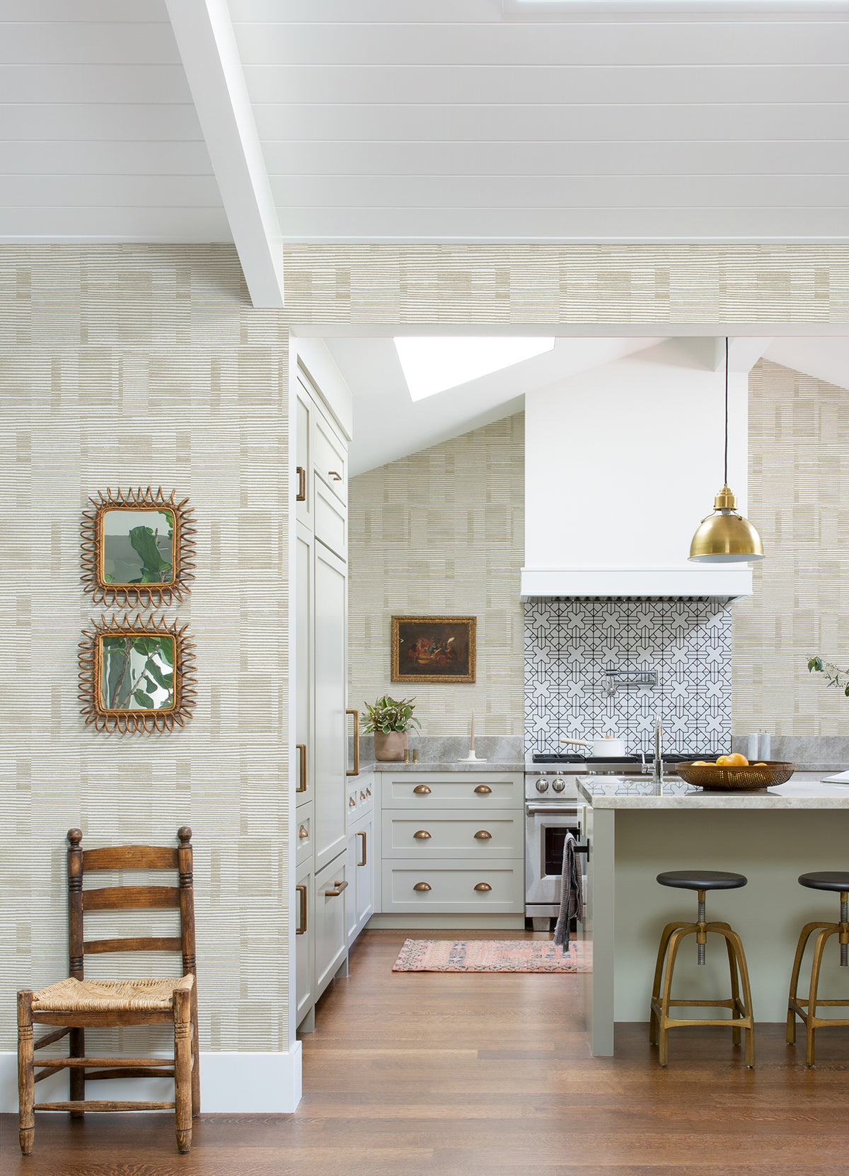 A bright kitchen features light cabinets, a patterned Callaway Woven Stripes Wallpaper by York Wallcoverings as the backsplash, a gold pendant light, two bar stools, decorative mirrors on the wall, and a wooden chair on a hardwood floor.