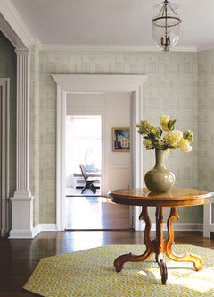 In a bright hallway adorned with York Wallcoverings’ Callaway Woven Stripes Wallpaper, a round wooden table topped with a green vase of yellow flowers sits on a yellow rug accented by white trim and columns. Through an open doorway, another room featuring a chair and artwork can be seen.