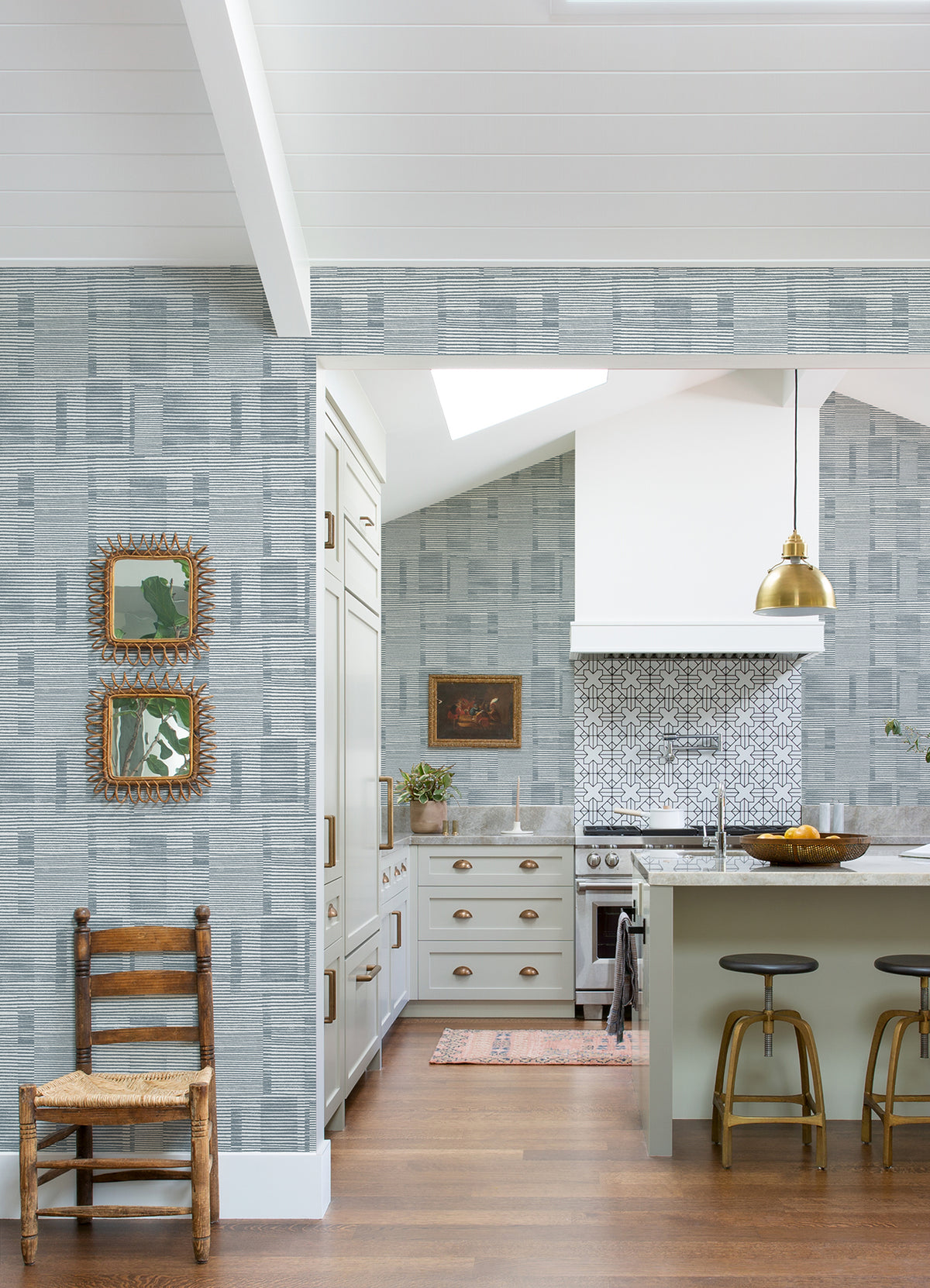 Bright kitchen featuring York Wallcoverings’ Callaway Woven Stripes Wallpaper, gold pendant lights, barstools at a green island, a wooden chair, and two framed mirrors on the wall. White cabinets, a patterned backsplash, and wooden floors complete the cozy space.