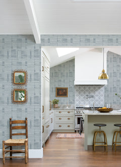 A modern kitchen features Callaway Woven Stripes Wallpaper by York Wallcoverings, a white stove, gold pendant light, three barstools, and wooden accents. Two framed mirrors and a wooden chair are set against the wall in the foreground.