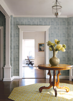In the hallway, a round wooden table topped with a green vase of yellow flowers sits on a yellow patterned rug. The walls are adorned with Callaway Woven Stripes Wallpaper by York Wallcoverings, complemented by white trim, columns, and a hanging glass light fixture. A bright room is visible through the doorway.
