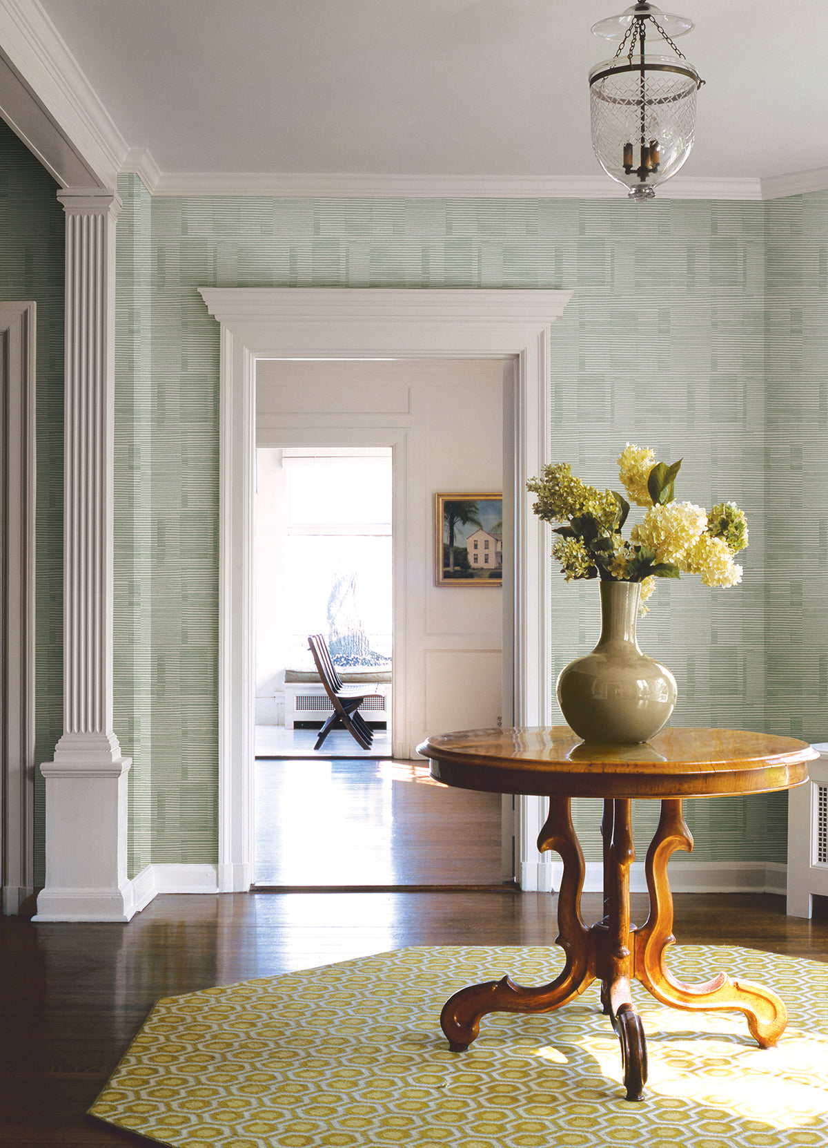A round wooden table topped with a green vase of yellow flowers sits on a yellow patterned rug in a bright room featuring York Wallcoverings’ Callaway Woven Stripes Wallpaper, complemented by white trim and columns. A doorway leads to another room furnished with a chair and a framed picture.