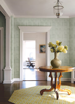 A round wooden table topped with a green vase of yellow flowers sits on a yellow patterned rug in a bright room featuring York Wallcoverings’ Callaway Woven Stripes Wallpaper, complemented by white trim and columns. A doorway leads to another room furnished with a chair and a framed picture.