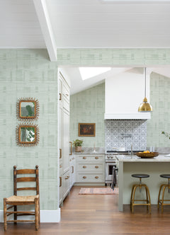 The kitchen features airy, high white ceilings and wood floors, with a bright atmosphere. The walls are adorned with Callaway Woven Stripes Wallpaper by York Wallcoverings in green patterns. The space includes a wooden chair, two mirrors, gold light fixtures, a white stove, an island with stools, and a patterned backsplash.