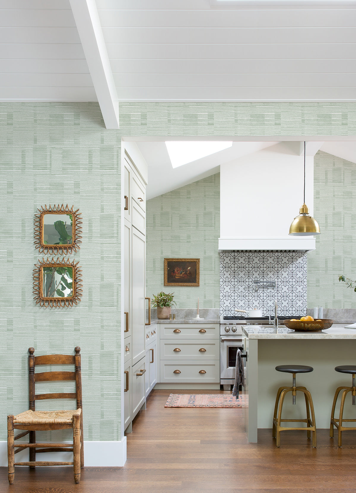 Bright kitchen featuring Callaway Woven Stripes Wallpaper by York Wallcoverings in green, complemented by white cabinets, a gold pendant light, patterned backsplash, wooden stools, a rustic chair, and two framed mirrors on the wall. Natural light fills the space.