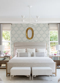 A modern, elegant bedroom featuring a large beige bed, two benches at the foot, matching nightstands with lamps, a gold oval mirror, and walls adorned with Zaria Topiary Wallpaper by York Wallcoverings. Two windows let in abundant natural light.