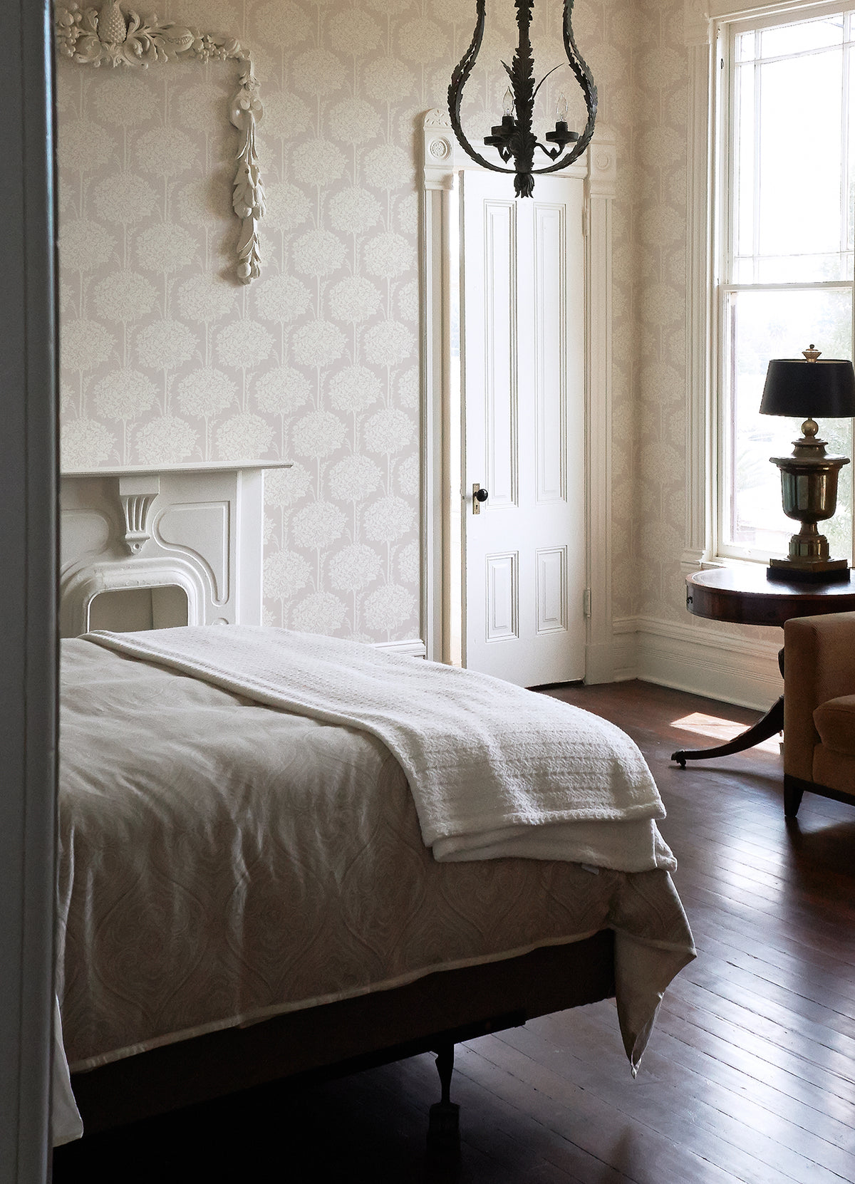 A sunlit bedroom featuring a neatly made bed, vintage chandelier, white ornate fireplace, round table with a lamp, and a large window allowing in natural light, with the walls adorned in York Wallcoverings’ Zaria Topiary Wallpaper.