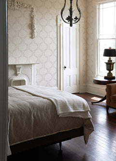A sunlit bedroom featuring a neatly made bed, vintage chandelier, white ornate fireplace, round table with a lamp, and a large window allowing in natural light, with the walls adorned in York Wallcoverings’ Zaria Topiary Wallpaper.