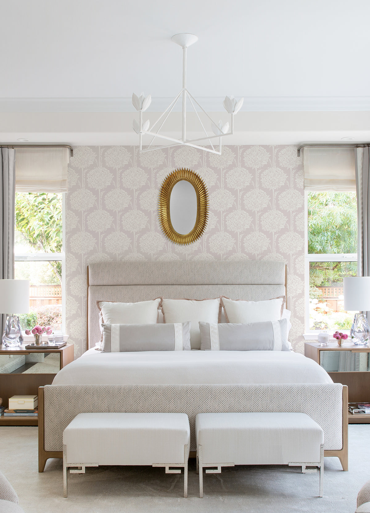 The bright bedroom features a king bed with light bedding, two benches at the foot, matching nightstands each adorned with lamps and flowers, two windows dressed in drapes, and a gold-framed oval mirror above the bed—all set against elegant Zaria Topiary Wallpaper by York Wallcoverings.