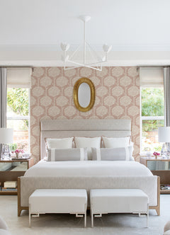 Transformed sentence using product data: Elegant bedroom featuring a large bed, two ottomans, nightstands with lamps, a gold oval mirror above the headboard, Zaria Topiary Wallpaper by York Wallcoverings adding a floral touch to the walls, and windows on both sides allowing natural light to fill the space.