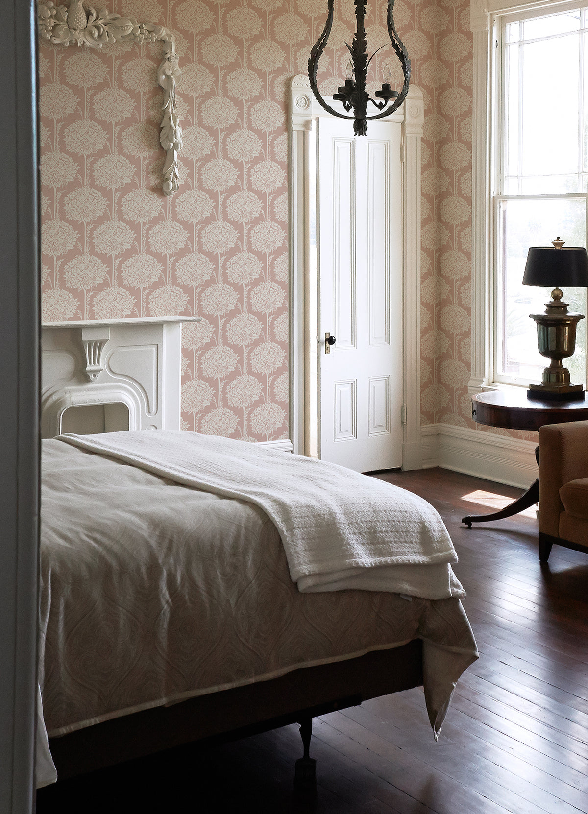 A sunlit bedroom with a large bed dressed in white bedding, a fireplace, chandelier, and a lamp on a round table near a tall window with sheer curtains features ornate Zaria Topiary Wallpaper by York Wallcoverings.