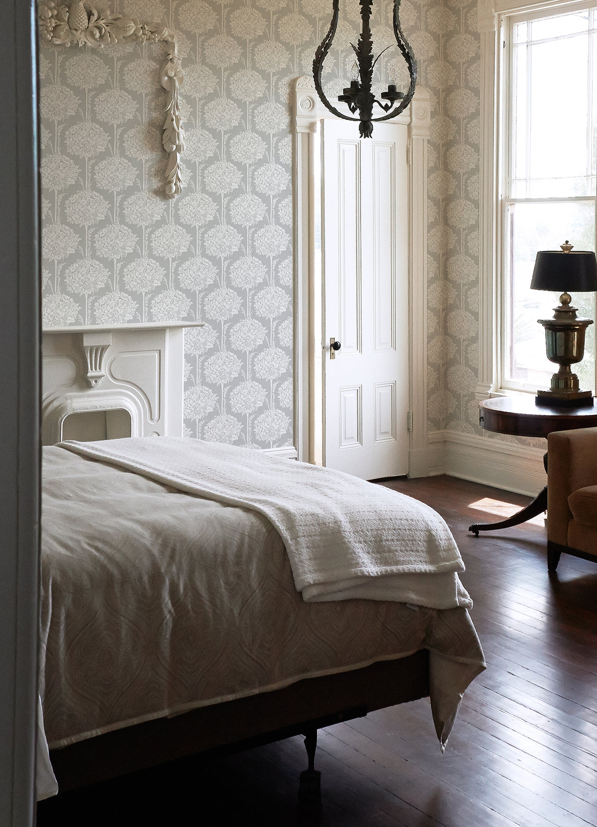 A bright bedroom features Zaria Topiary Wallpaper by York Wallcoverings, a white bed with a blanket, a decorative fireplace, a chandelier, wooden floors, a round table with a lamp, an armchair, and large windows filled with natural light.