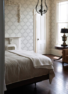 A bright bedroom features Zaria Topiary Wallpaper by York Wallcoverings, a white bed with a blanket, a decorative fireplace, a chandelier, wooden floors, a round table with a lamp, an armchair, and large windows filled with natural light.