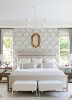 Bright bedroom featuring a large upholstered bed, two benches at the foot, matching nightstands with lamps, Zaria Topiary Wallpaper by York Wallcoverings adding a floral touch, a round gold mirror, and two windows with white curtains showcasing greenery outside.