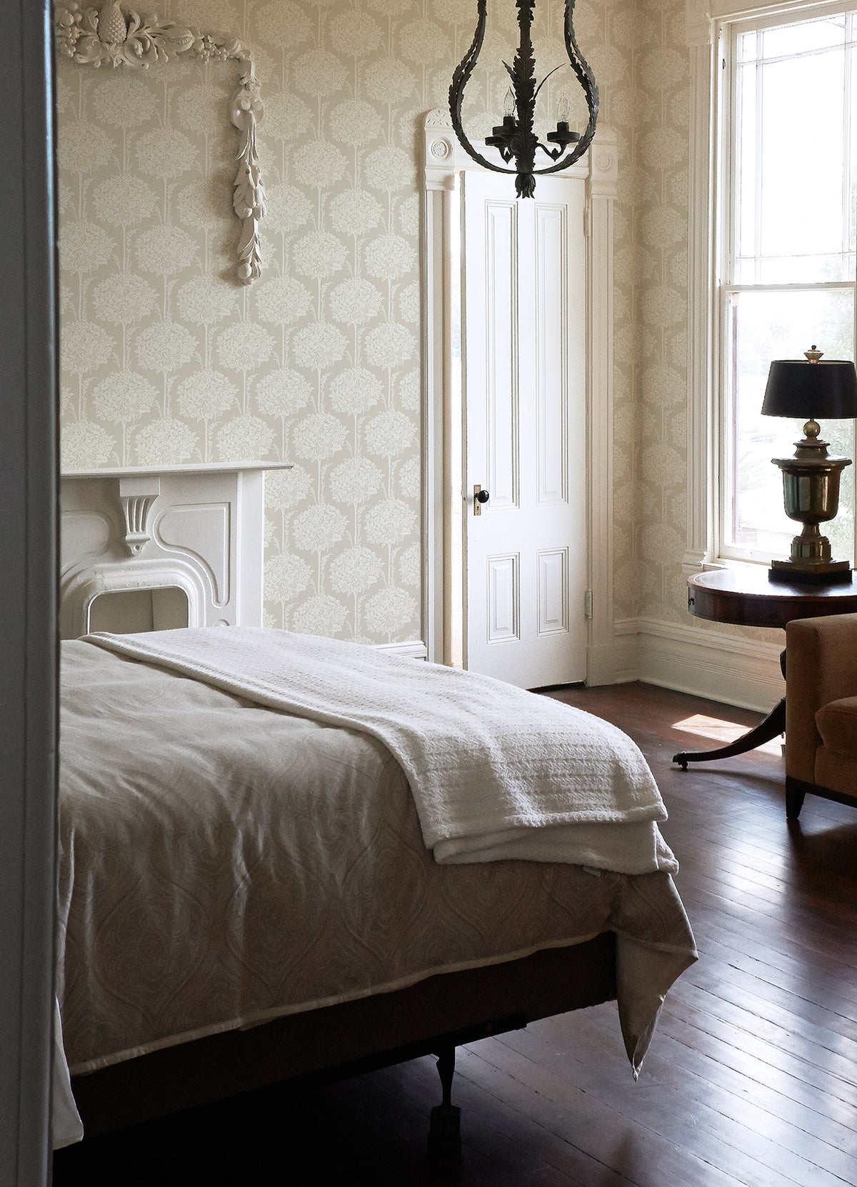 A bright bedroom features a white bed, a decorative fireplace, tall windows streaming in natural light, a chandelier, an armchair, a round table with a lamp, and York Wallcoverings’ Zaria Topiary Wallpaper adding patterned elegance to the space.