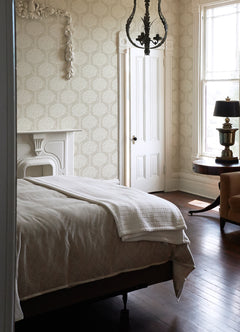 A bright bedroom features a white bed, a decorative fireplace, tall windows streaming in natural light, a chandelier, an armchair, a round table with a lamp, and York Wallcoverings’ Zaria Topiary Wallpaper adding patterned elegance to the space.