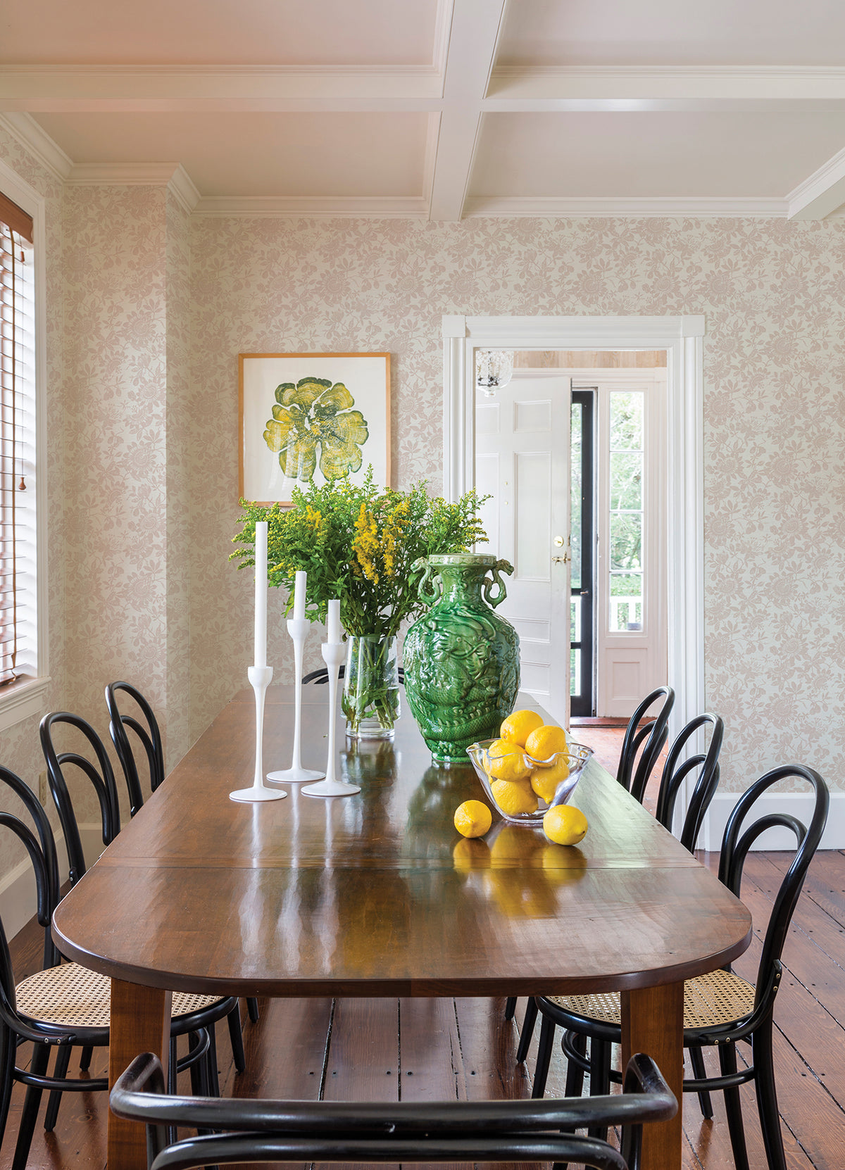 A wooden dining table with black chairs is set with white candlesticks, a green vase filled with yellow flowers, and a bowl of lemons. The bright dining room features Helen Floral Trail Wallpaper by York Wallcoverings and a framed artwork.